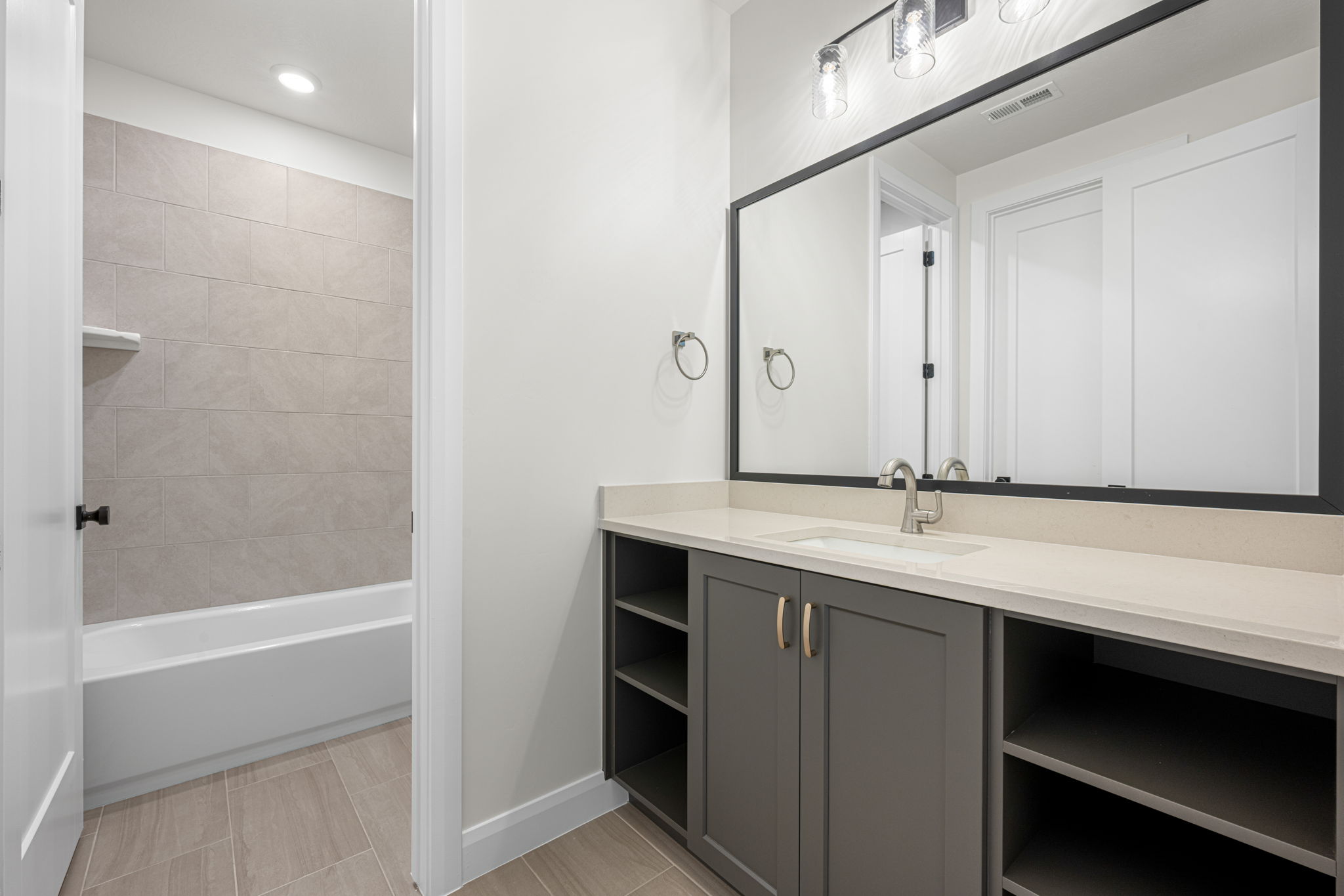 Bright bathroom featuring a spacious vanity with open shelving, black-framed mirror, and separate tiled bathtub area.