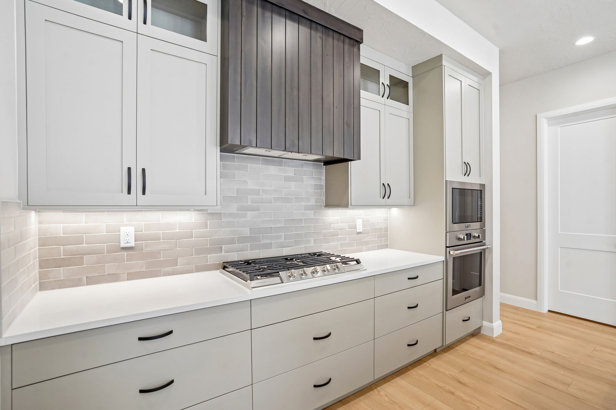Modern kitchen in the Pacific II by Cole West featuring custom gray cabinetry, a decorative wood range hood, tile backsplash, and built-in stainless steel appliances.