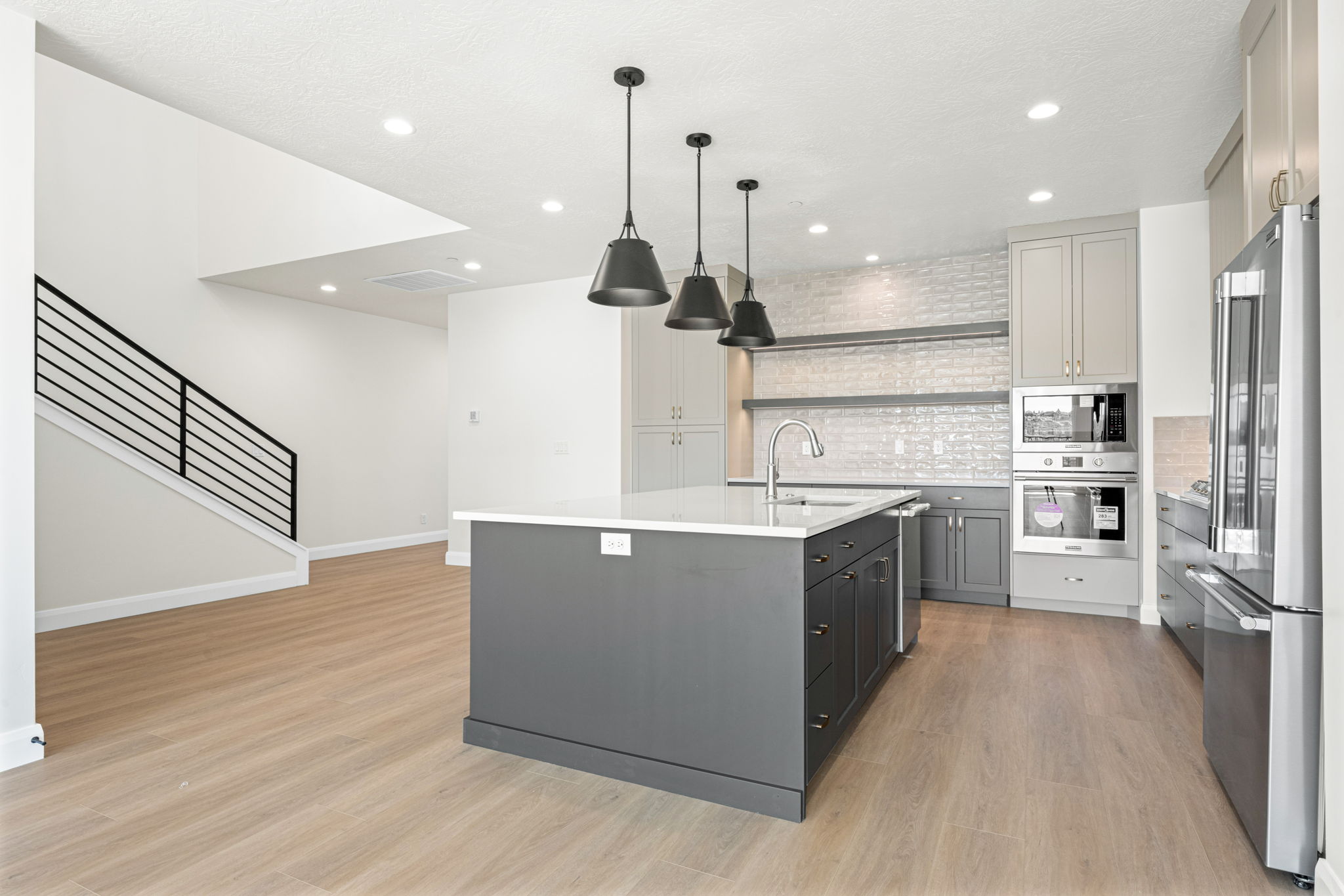 Spacious modern kitchen featuring a large gray island, white countertops, black pendant lighting, stainless steel appliances, and an open staircase.