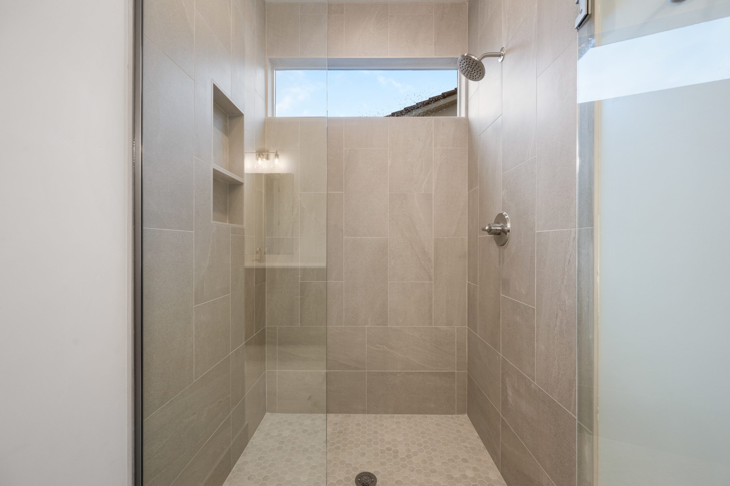 Modern walk-in shower in the Mesa primary bathroom at Solis, featuring floor-to-ceiling neutral tile, glass enclosure, built-in shelving, and a horizontal window.