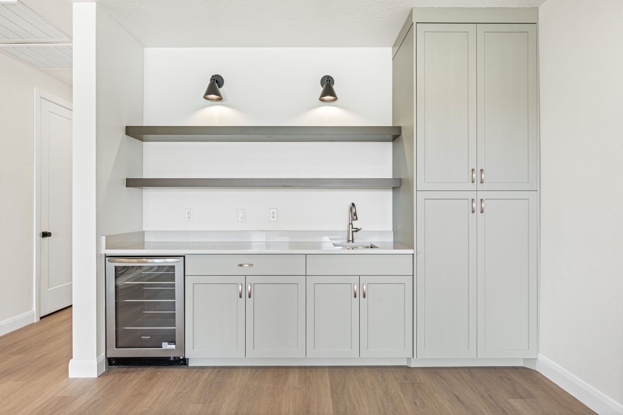 Built-in wet bar featuring gray cabinetry, floating shelves, beverage refrigerator, white countertop, and modern black wall sconces.